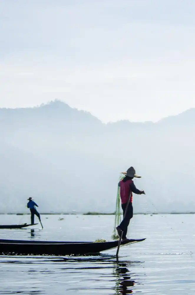Fisherman Casting Net from Traditional Boat on Misty Himalayan Lake, Pokhara Nepal.webp